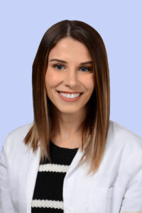 Headshot of Katie Shea, PA-C on a light blue background. Katie is smiling and wearing a black and white striped shirt under a white coat.